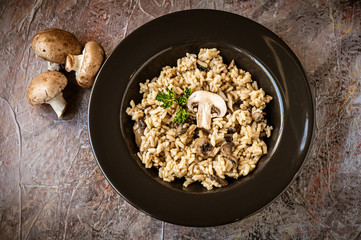 Top down view of a bowl of mushroom risotto next to three brown mushrooms in a black bowl on a  textured surface