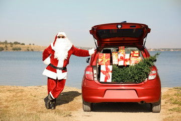 Authentic Santa Claus near car with open trunk full of presents and fir tree outdoors
