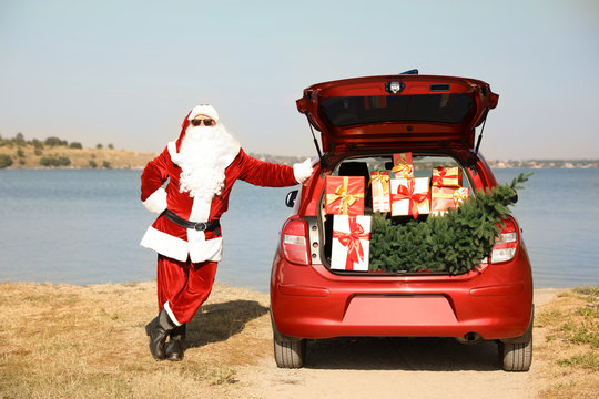 Authentic Santa Claus Near Car With Open Trunk Full Of Presents And Fir Tree Outdoors