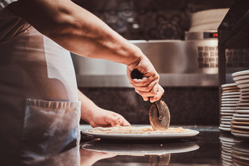 Chef is cutting tasty pizza with special knife at the kitchen.