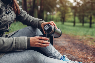 Fototapeta premium Woman tourist pours hot tea out of thermos in autumn forest. Camping, traveling and sport concept