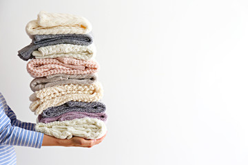 Slim young woman wearing oversized cotton shirt with blue stripes & holding big stack of folded knitted warm pastel color sweaters, easy chic style, different knitting patterns. Background, copy space