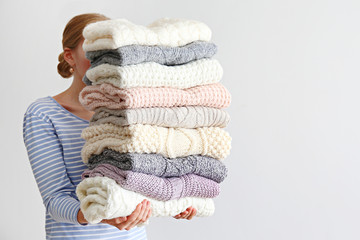 Slim young woman wearing oversized cotton shirt with blue stripes & holding big stack of folded knitted warm pastel color sweaters, easy chic style, different knitting patterns. Background, copy space