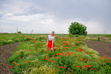 Blonde young woman in red skirt and white shirt, red earrings is in the middle of a poppy field.