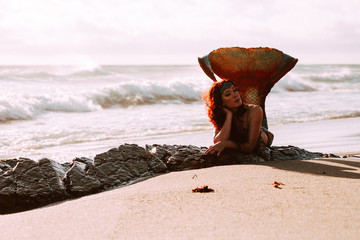 Caucasian redhead woman with mermaid tail reclines on the beach in the waves
