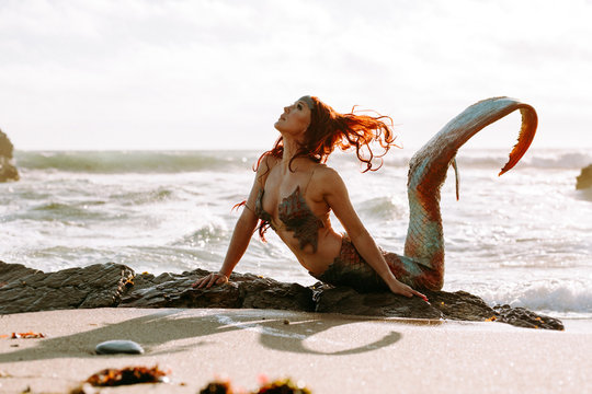 Caucasian redhead woman with mermaid tail arches her back in the surf at sunset