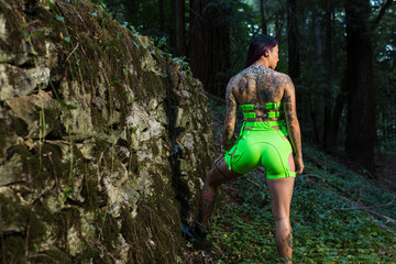 Asian tattooed woman in neon rave clothing poses on an abandoned rock wall in the forest