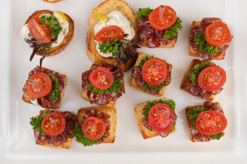 Close-up of wedding reception with tasty sandwiches and canapes made of sausage, parsley and tomatoes with cream cheese. Canapes on white ceramic plates at wedding reception.