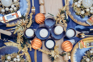 Top view Christmastime table setting, festive dinnerware decorated with details and candles in Blue and Gold colors. Navy Blue Table Linens and Gold Cutlery. Trends of Winter Holiday Tablescape Decor.