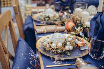 Christmastime table setting, festive dinnerware decorated with details and white balls in Blue and Gold colors. Navy Blue Table Linens and Gold Cutlery. Trends of Winter Holiday Tablescape Decor.