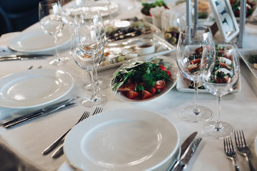 View over wedding table covered with white tablecloth with empty glasses, plates and utensils. Snacks are served on the table.