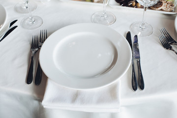 Close-up empty serving white big plate surrounded by knife fork ready to eating food high angle. Beautiful tableware on white tablecloth at luxury fashionable restaurant before dinner