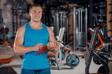 Fotobehang Persoonlijk Portrait of a man, a personal trainer holding a clipboard with a training plan in the gym.  © fusssergei