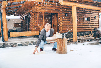 Man chopping wood on snowy yard for a house fireplace with heavy snowflakes background . Winter countryside holidays concept image