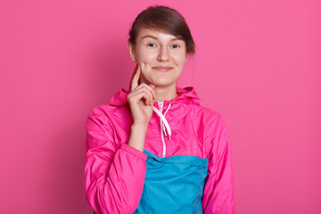 Portrait of smiling young woman looking directly at camera, standing isolated over pink background, attractive female wearing sporty blue rose shirt, keep sher index finger on cheek. People concept.