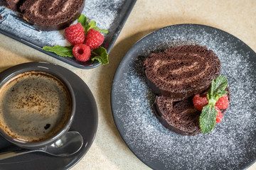 Chocolate swiss roll decorated with raspberries on a plate and black coffee on the table. Selective focus.