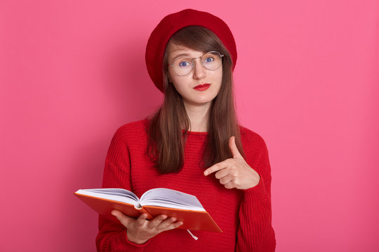 Horizontal shot of woman with orange note book pointing at its pages with her index finger. Attractive female wearing red sweater, beret and glasses, having perfect skin and bright pomade makeup.