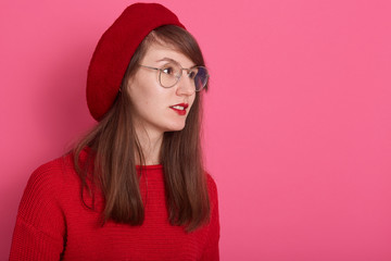 Close up portrait of pensive young female dresses sweater, beret and round spectacles, looking thoughtfully aside, standing against rosy studio wall, copy space for advertisment or promotion.