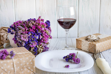 Table setting. Plate on wooden background with gift box, cutlery, a glass of red wine and flowers.