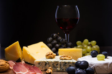 Assorted cheeses, nuts, grapes, fruits, smoked meat and a glass of wine on a serving table. Dark and Moody style.
