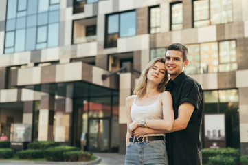 Fototapeta premium Positive Young family stands on the street against the backdrop of a modern apartment building,hugging and looking camera, wearing casual clothes.Street portrait of happy couple posing at camera