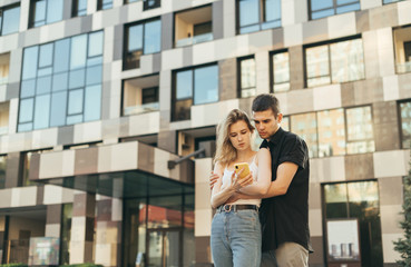 Beautiful young couple in stylish casual clothes stands on the background of modern architecture, hugging and browsing the internet on a smartphone, looking at the screen with serious faces.