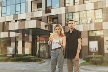 Fototapeta premium Street portrait of a fun couple in stylish casual clothes standing on the street and looking into the camera against the background of a modern apartment building.
