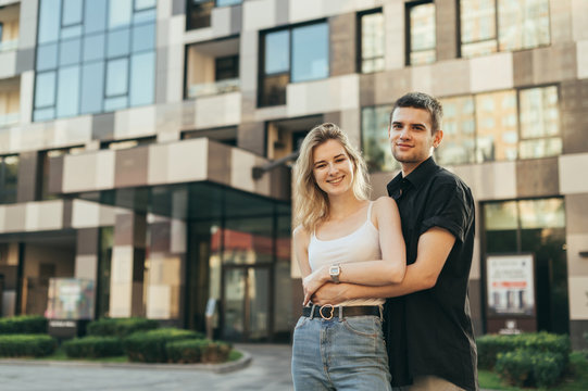 Street Photo Beautiful Stylish Young Couple Hugging Against The Backdrop Of A Modern Building, Looking In Camera And Smiling. Happy Young Family Stands On The Background Of A Modern Apartment Building