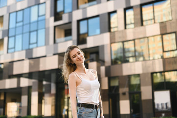 Fototapeta na wymiar Smiling girl in a white T-shirt and jeans is standing outside, looking at the camera and smiling. Street portrait of stylish positive girl in casual clothes on a walk