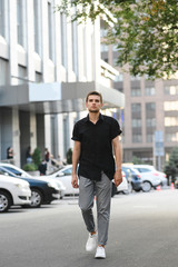 Full length portrait of a young man in stylish casual clothes, wearing a black shirt and gray pants, walking down the city street and looking into the camera. Vertical photo.