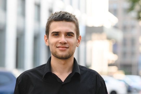 Happy young man in black shirt on urban background looking into camera and smiling. Closeup portrait of handsome guy in dark shirt on light urban background.