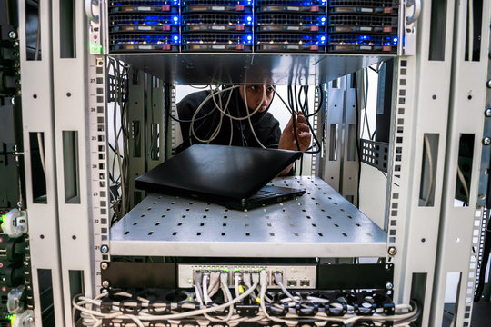 Hacker switches the wires in the server room. The malefactor is behind the computer equipment of the data center. A man in a mask unravels communication cable internet.