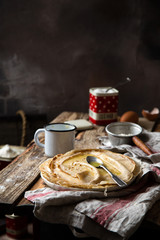 traditional russian or ukrainian stack of thin crepes with spoon of melted butter on top stands on rustic wooden table with vintage cup, strainer opposite concrete grey wall