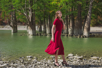a beautiful, young brown-haired woman in a red dress on the background of a morning lake with cypresses, a girl with a slender figure posing against nature