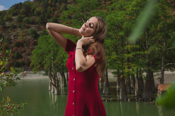 a beautiful, young brown-haired woman in a red dress on the background of a morning lake with cypresses, a girl with a slender figure posing against nature