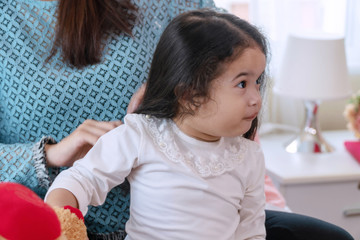 Little cute girl sitting on the bed while her mother combing daughter's hair together. Cheerful adorable childhood feel comfort with mom making hairstyle at home while looking at something.