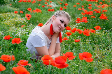 Blonde young woman in red skirt and white shirt, red earrings is in the middle of a poppy field.