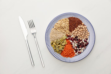 top view of fork and knife near striped plate with raw lentil, chickpea, quinoa, oatmeal, beans and pumpkin seeds on marble surface