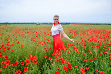 Blonde young woman in red skirt and white shirt, red earrings is in the middle of a poppy field.