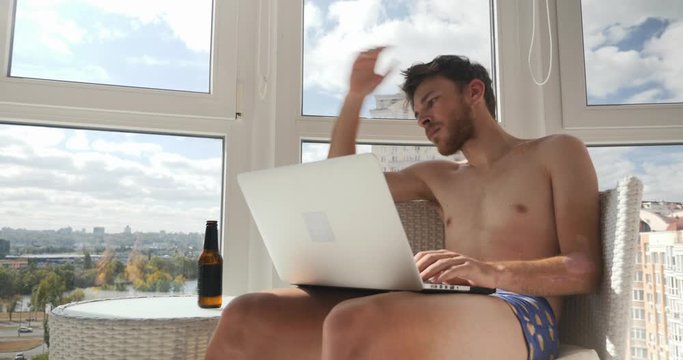 Arrogant pathos guy in underpants sitting at modern luxury balcony, playing video games and drinking beer. Young drunk male person looking at monitor and drinking alcohol