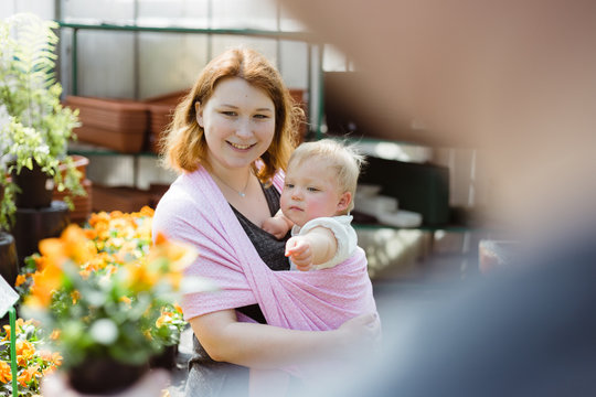 Close up of mother carrying her baby daughter in a pink sling wrap while shopping for flowers for their garden in a local greenhouse - Powered by Adobe