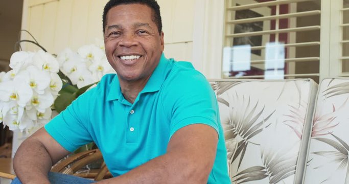 Portrait of attractive older African American man smiling while sitting on porch looking at camera. Happy and handsome black man in his 60s on a sunny day. Slow motion 4k handheld