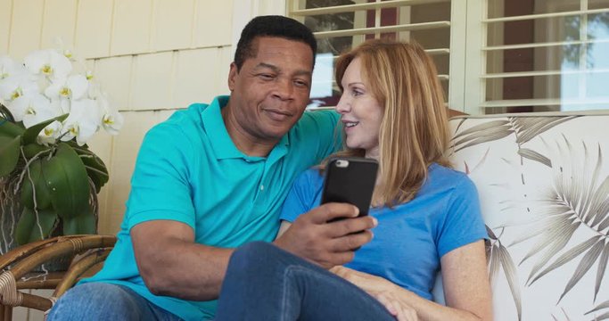 Happy older couple sitting on porch and using smartphone together. African American and Caucasian husband and wife looking at cell phone on sunny day outside home. Slow motion 4k handheld