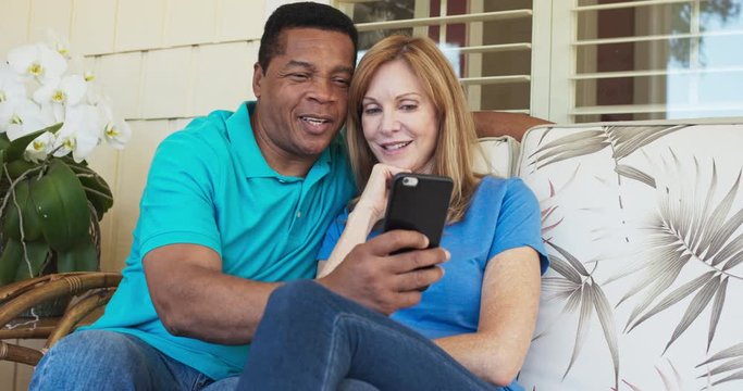 Happy older couple sitting on porch and using cell phone together. African American and Caucasian husband and wife looking at smartphone on sunny day outside home. Slow motion 4k handheld