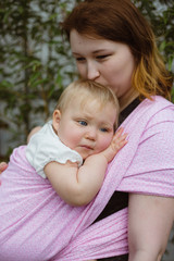 Young redhead babywearing mother kissing her sleepy one year old baby girl wrapped in a pink sling 