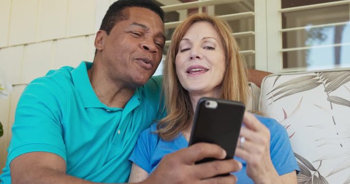 Close up of Smiling older couple sitting on porch and using cellphone together. African American and Caucasian husband and wife looking at mobile phone at home. Slow motion 4k handheld