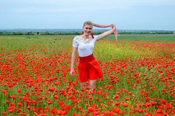 Blonde young woman in red skirt and white shirt, red earrings is in the middle of a poppy field.