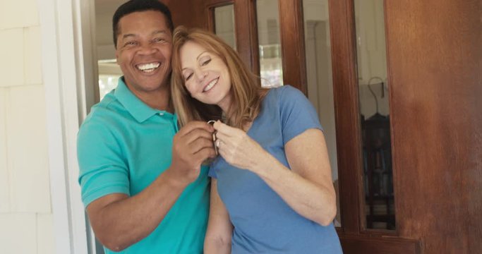 Happy older couple holding the keys to their new house while standing at the front door. African American and Caucasian husband and wife homeowners purchase a home. Slow motion 4k handheld