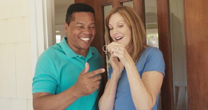Happy mature couple holding the keys to their new house while standing at the front door. African American and Caucasian husband and wife homeowners purchase a home. Slow motion 4k handheld