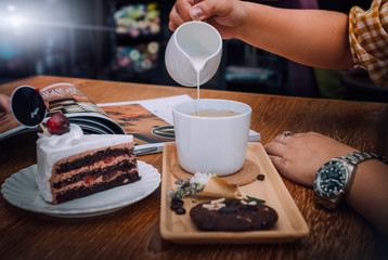 Woman holding milk cream mug for filling in the coffee on wooden table, Which her breakfastwill consist of blueberry jam on ceramic plate and chocolate chip cookie with hot coffee.